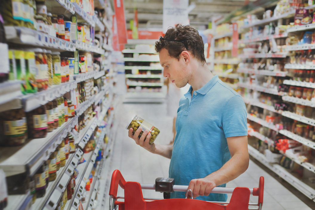 man buying food in supermarket reading ingredients on a jar of olives