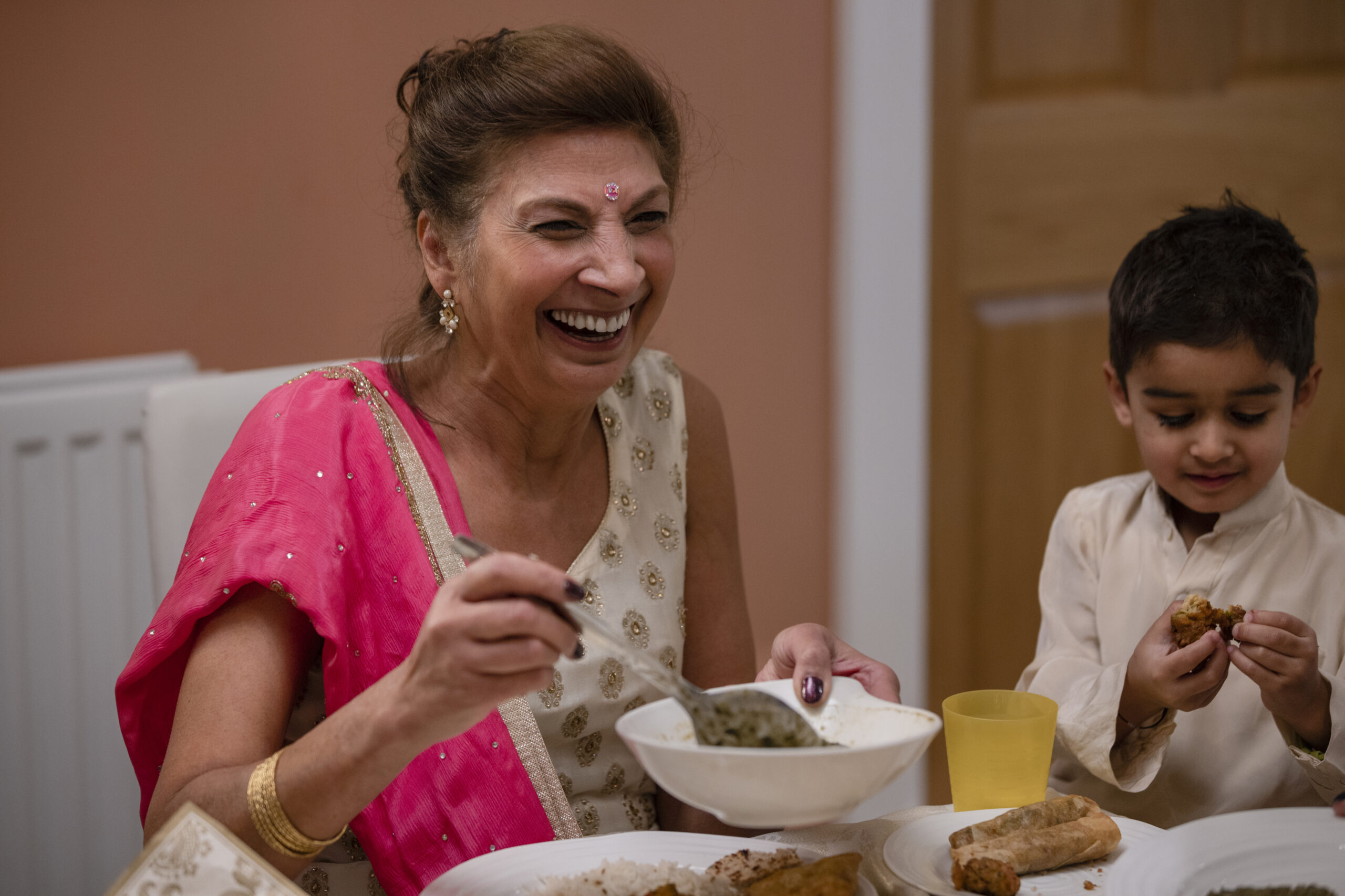 A close-up front view of a cheerful happy grandmother sitting next to her grandson as they sit down for a family meal to celebrate Diwali as a family by having lots of tasty home-cooked traditional foods.